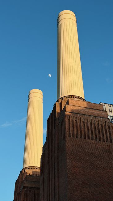 A large brick building with four tall, cylindrical white chimneys rising from its roof, situated beside a body of water. The building appears to be a historic power station, with prominent brickwork a