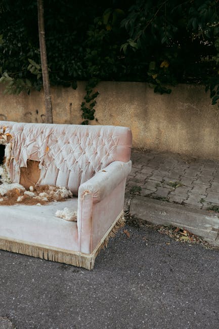 A light pink upholstered sofa with a tufted backrest and decorative fringes along the base is positioned outdoors on a paved area near a concrete wall and a pavement. The sofa's fabric appears worn an