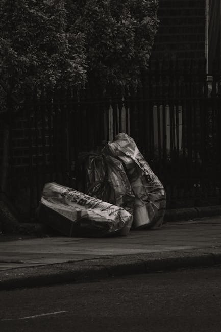 A light pink upholstered sofa with a tufted backrest and decorative fringes along the base is positioned outdoors on a paved area near a concrete wall and a pavement. The sofa's fabric appears worn an
